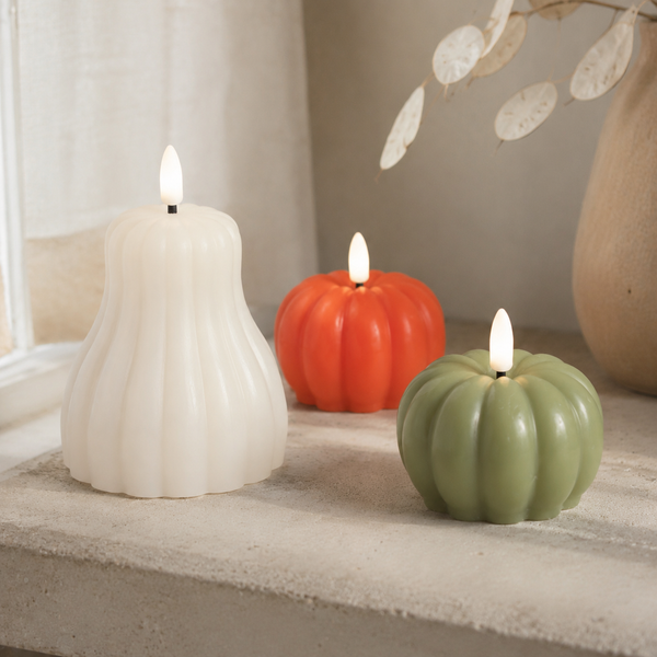 Three flameless LED pumpkin-shaped candles in white, orange, and sage green, styled on a stone windowsill with a neutral background.