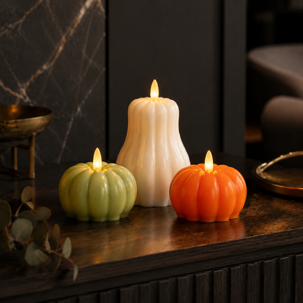 Three decorative electric pumpkin candles in green, white, and orange, lit and arranged on a dark wooden mantelpiece or console table against a dark marble wall.