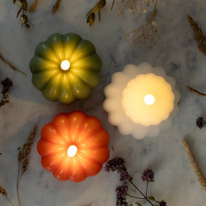 Three decorative pumpkin-shaped LED tea lights glowing on a light marble surface with dried flowers and wheat scattered around them. One tea light is olive green, one is orange, and one is frosted white.