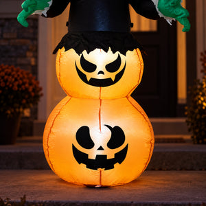 A glowing inflatable Halloween decoration shaped like a witch's broom or broom-style jack-o'-lantern stack, with two orange pumpkin forms, black facial cutouts, a black hat, green gloved hands, and a dark doorway background.