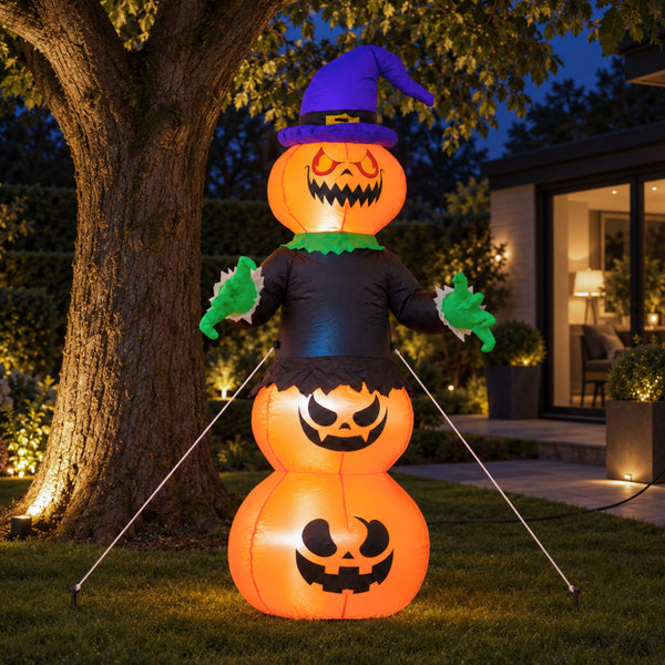 An inflatable Halloween yard decoration shaped like a witch jack-o'-lantern, standing on grass in front of a house at night. The figure has three stacked glowing orange pumpkin bodies, a purple witch hat, a black dress, green gloves, and green collar details.