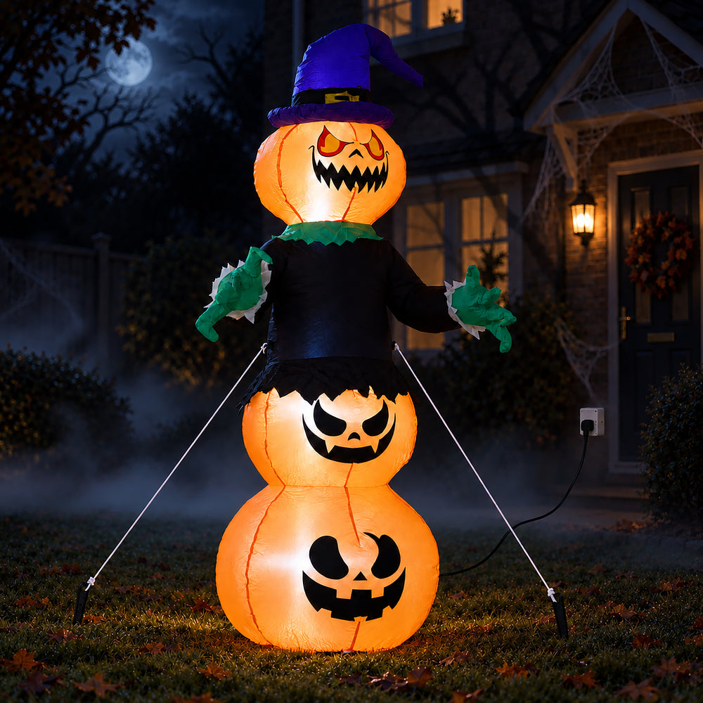A Halloween inflatable jack-o'-lantern stack with a witch hat and black outfit, photographed outdoors in front of a house at night on a lawn.