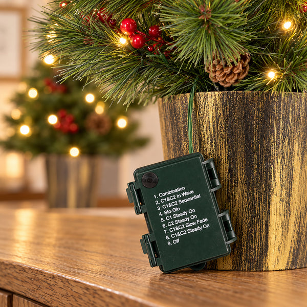 A small dark green electronic control box with printed text labels hangs from a green string beneath a decorated Christmas tree in a rustic wooden planter, with a softly blurred Christmas tree in the background.