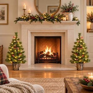 A cozy living room scene centered on a lit fireplace with a white stone mantel decorated with green garland, candles, pinecones, and warm lights. Two small decorated Christmas trees stand on either side of the fireplace, and a sofa and coffee table are partially visible in the foreground.