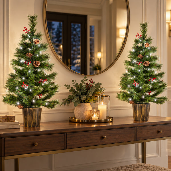 Two small decorated Christmas trees with white fairy lights and red berry ornaments stand on a wooden console table in a warmly lit room, with a mirror and candles in the background.