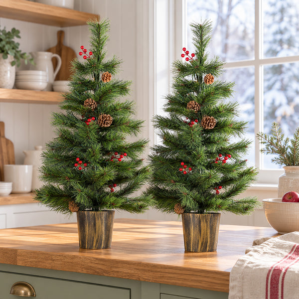 Two small artificial Christmas trees with green needles, brown pinecones, and red berries, displayed on a wooden kitchen counter in front of a window.