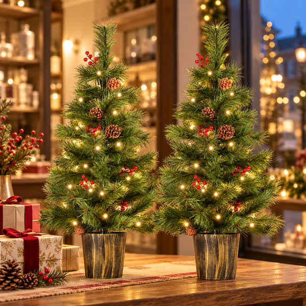 Two small artificial Christmas trees with warm white lights, red berries, and pinecones, displayed on a wooden table in a cozy holiday shop setting with wrapped gifts and blurred lights in the background.
