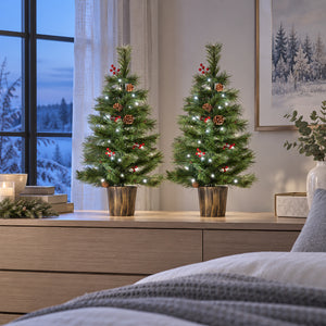 Two small artificial Christmas trees with white string lights, pinecones, and red berry ornaments, displayed on a wooden dresser in a cozy bedroom setting.