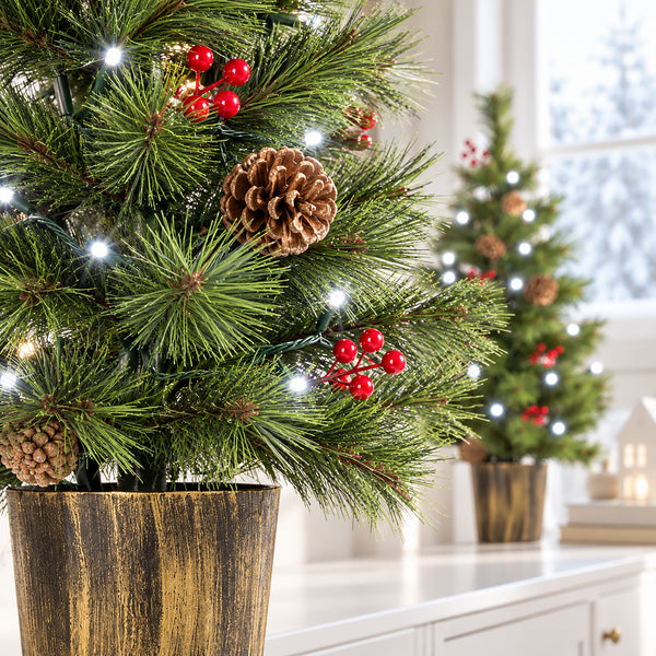 A close-up of a potted artificial Christmas tree with green pine needles, pinecones, red berries, and glowing white string lights in a rustic gold pot, with a softly blurred second tree in the background.