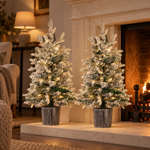 Set of two 90cm pre lit potted Christmas trees with snowy branches and warm white LED lights beside a fireplace.