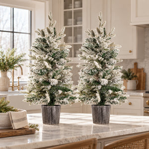 Two frosted Christmas trees in dark gray metal planters, displayed on a marble kitchen island with a bright, modern kitchen in the background.