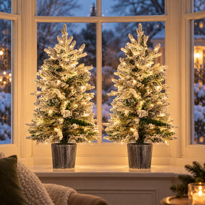 Two small pre lit snowy Christmas trees with warm white LED lights displayed on a festive windowsill.