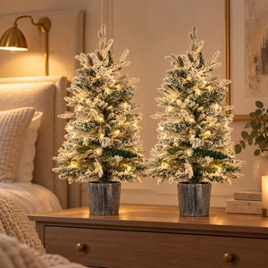 Set of two pre lit snow flocked Christmas trees with warm white lights displayed on a bedroom sideboard.