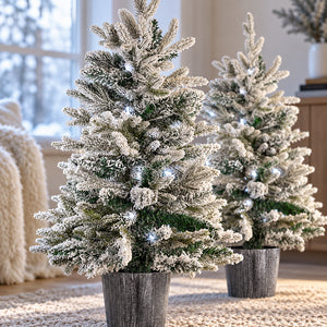 Two flocked Christmas trees with white snow-dusted needles and small warm white string lights, standing in gray faux-wood planters on a woven rug in a softly lit indoor setting.