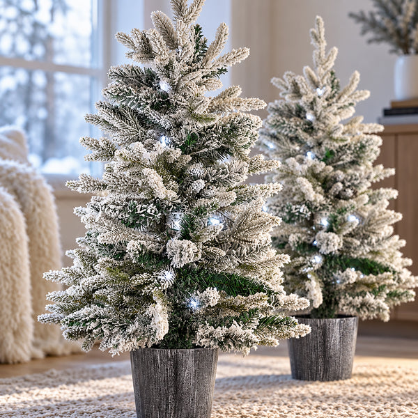 Two flocked Christmas trees with white snow-dusted needles and small warm white string lights, standing in gray faux-wood planters on a woven rug in a softly lit indoor setting.