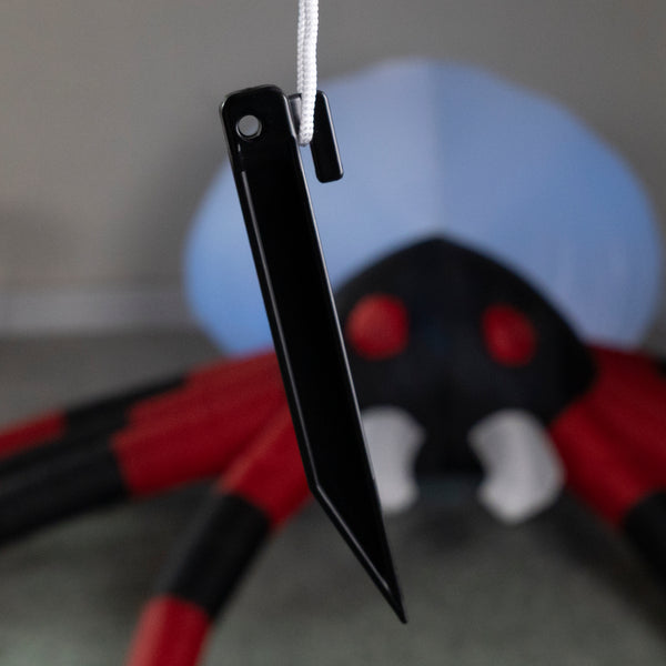 A black plastic ninja-style throwing star with a white cord, shown in sharp focus against a blurred background of a red and black spider decoration.