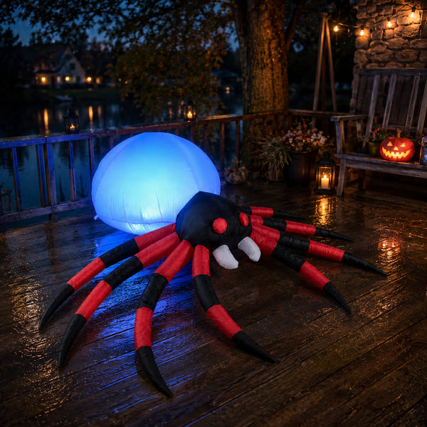 A large inflatable spider decoration with red and black striped legs and white fangs sits on a wet wooden deck at dusk, with a glowing blue orb behind it and a porch setting in the background.