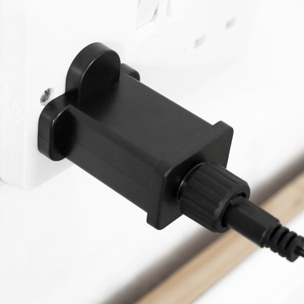 A close-up of a black plastic electrical adapter or plug inserted into a white wall outlet, with part of the attached cord visible.