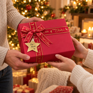 A red square gift box wrapped with a red ribbon and bow, held by two people in a cozy Christmas setting. The ribbon has gold lettering and a gold star-shaped tag that says 'For Mum'.