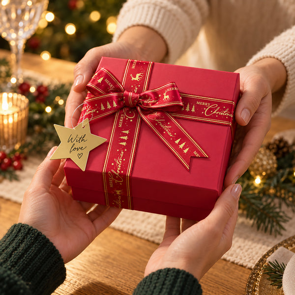 A red square gift box wrapped with a red ribbon printed with gold Christmas motifs and a gold star tag reading 'With love', held by two hands in a cozy holiday setting with warm lights and pine greenery in the background.