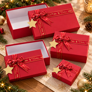 A set of red Christmas gift boxes in different sizes, decorated with red ribbons featuring gold lettering and gold star-shaped tags, arranged on a decorated Christmas table with pine branches and lights.