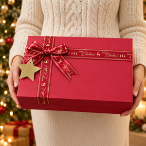 A person in a cream cable-knit sweater holds a red Christmas gift box tied with a red ribbon and bow, featuring a gold star ornament and gold holiday script details. A blurred Christmas tree with warm lights is in the background.