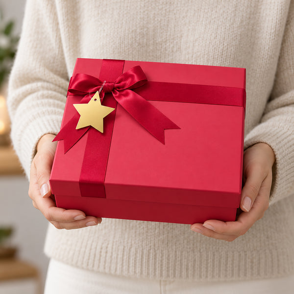 A person in a cream knit sweater holds a red gift box tied with a red ribbon and bow, decorated with a small gold star tag.