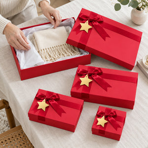 A set of red gift boxes with red ribbons and gold star ornaments, shown on a beige tablecloth. One open box reveals a cream-colored fringed scarf, and a person's hands are visible adjusting the tissue paper inside.