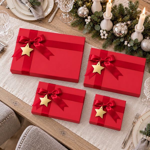 Four red gift boxes of different sizes arranged on a dining table, each tied with a red ribbon and bow and decorated with a gold star ornament. The scene includes holiday greenery, candles, plates, and glassware in the background.