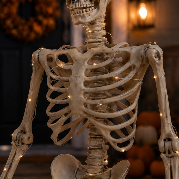 A Halloween skeleton decoration with warm white string lights draped over the rib cage and spine, shown in a dimly lit autumn-themed setting with pumpkins and a lantern in the background.