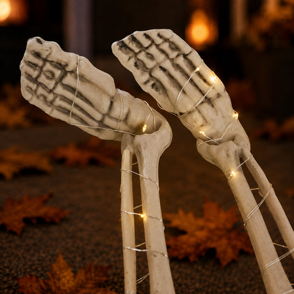 Two decorative skeleton hand props wrapped with warm white fairy lights, standing upright on a dark surface with scattered autumn leaves in a softly blurred setting.