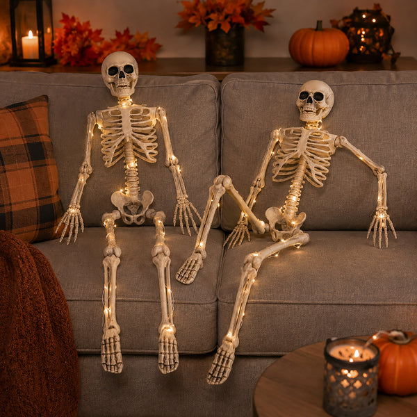Two decorative skeleton figures seated on a gray couch, lit with warm white string lights. The scene is styled for Halloween with pumpkins, fall leaves, candles, and a plaid pillow in a real indoor environment.