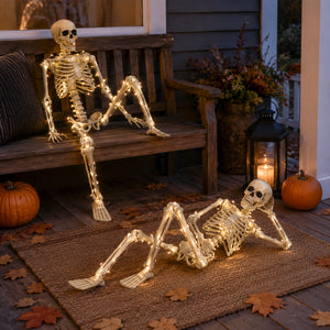 Two illuminated skeleton Halloween decorations posed on a rustic wooden porch. One skeleton sits on a wooden bench, and the other reclines on a woven rug on the deck.