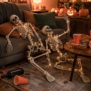 Two life-sized decorative skeletons with warm white string lights posed on a gray living room rug in front of a couch, with pumpkins and a cozy Halloween living room background.