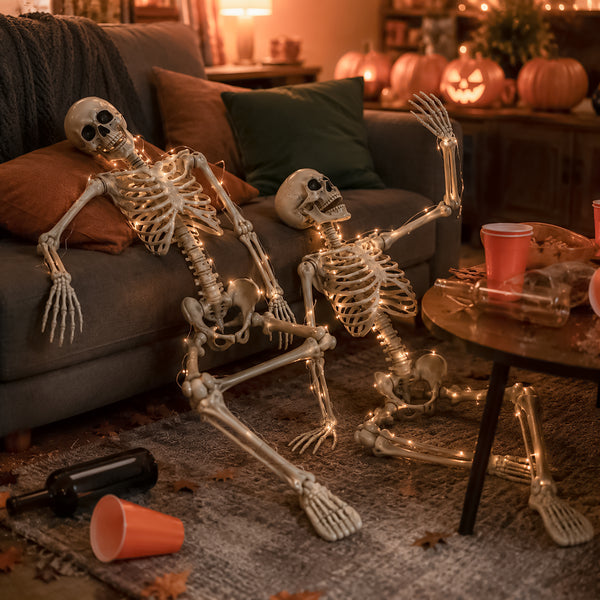 Two life-sized decorative skeletons with warm white string lights posed on a gray living room rug in front of a couch, with pumpkins and a cozy Halloween living room background.