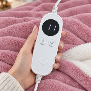 A hand holds a white digital remote control with a black display screen and buttons for temperature and time, placed over a pink plush blanket.