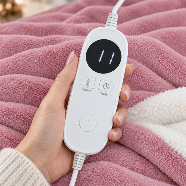 A hand holds a white digital remote control with a black display screen and buttons for temperature and time, placed over a pink plush blanket.