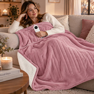 A woman reclines on a beige sofa in a cozy living room, wrapped in a pink fleece blanket with a white sherpa-style reverse side. She holds a white handheld controller, suggesting a heated blanket.