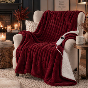 A plush electric heated throw blanket in deep burgundy red with a white sherpa lining, draped over a cream armchair in a cozy living room setting with a fireplace, candles, and a side table.
