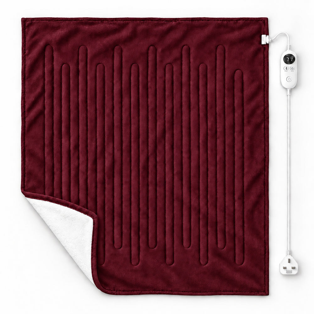 A plush electric heated throw blanket in deep burgundy red with a white sherpa lining, draped over a cream armchair in a cozy living room setting with a fireplace, candles, and a side table.