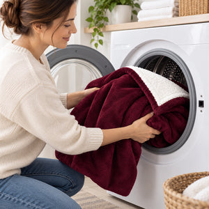 A woman in a cream sweater and blue jeans loads a burgundy fleece blanket with a white sherpa lining into a white front-loading washing machine in a bright, modern laundry room.