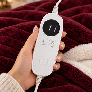 A hand holds a white electric blanket controller with a digital display showing '11' and buttons for temperature and time, resting over a plush burgundy fleece blanket with a cream sherpa lining.