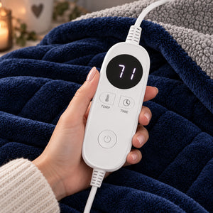 A hand holds a white remote control for an electric heated blanket, which displays a temperature of 71 degrees on its screen. The remote is positioned over a cozy, dark blue plush blanket with a gray sherpa lining.