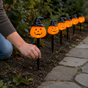 A person's hand is shown planting a row of glowing orange Halloween jack-o'-lantern yard lights with black witch hats on stakes in a garden bed next to a stone path.