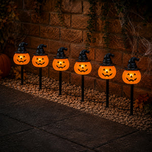 Six Halloween yard lights shaped like glowing jack-o'-lantern pumpkins with black witch hats, mounted on black stakes along a stone path in front of a brick wall.