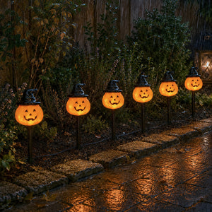 Five glowing jack-o'-lantern Halloween yard lights with witch hats on black stakes, arranged along a stone path in a rainy garden setting.