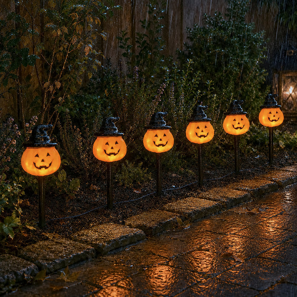 Five glowing jack-o'-lantern Halloween yard lights with witch hats on black stakes, arranged along a stone path in a rainy garden setting.