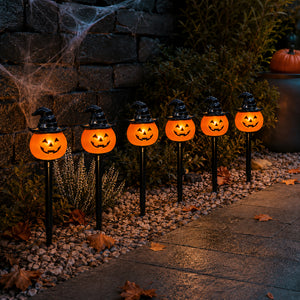 Six glowing jack-o'-lantern Halloween yard lights with witch hats are arranged along a stone path in a garden setting at night.