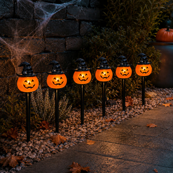 Six glowing jack-o'-lantern Halloween yard lights with witch hats are arranged along a stone path in a garden setting at night.