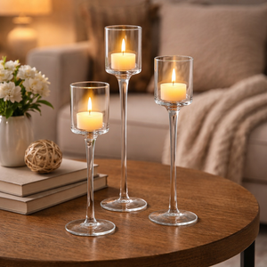 Three clear glass candle holders with long stems are arranged on a round wooden table in a cozy living room setting. Each holder contains a lit white tea light candle, with a vase of white flowers and stacked books on the left and a blurred sofa in the background.